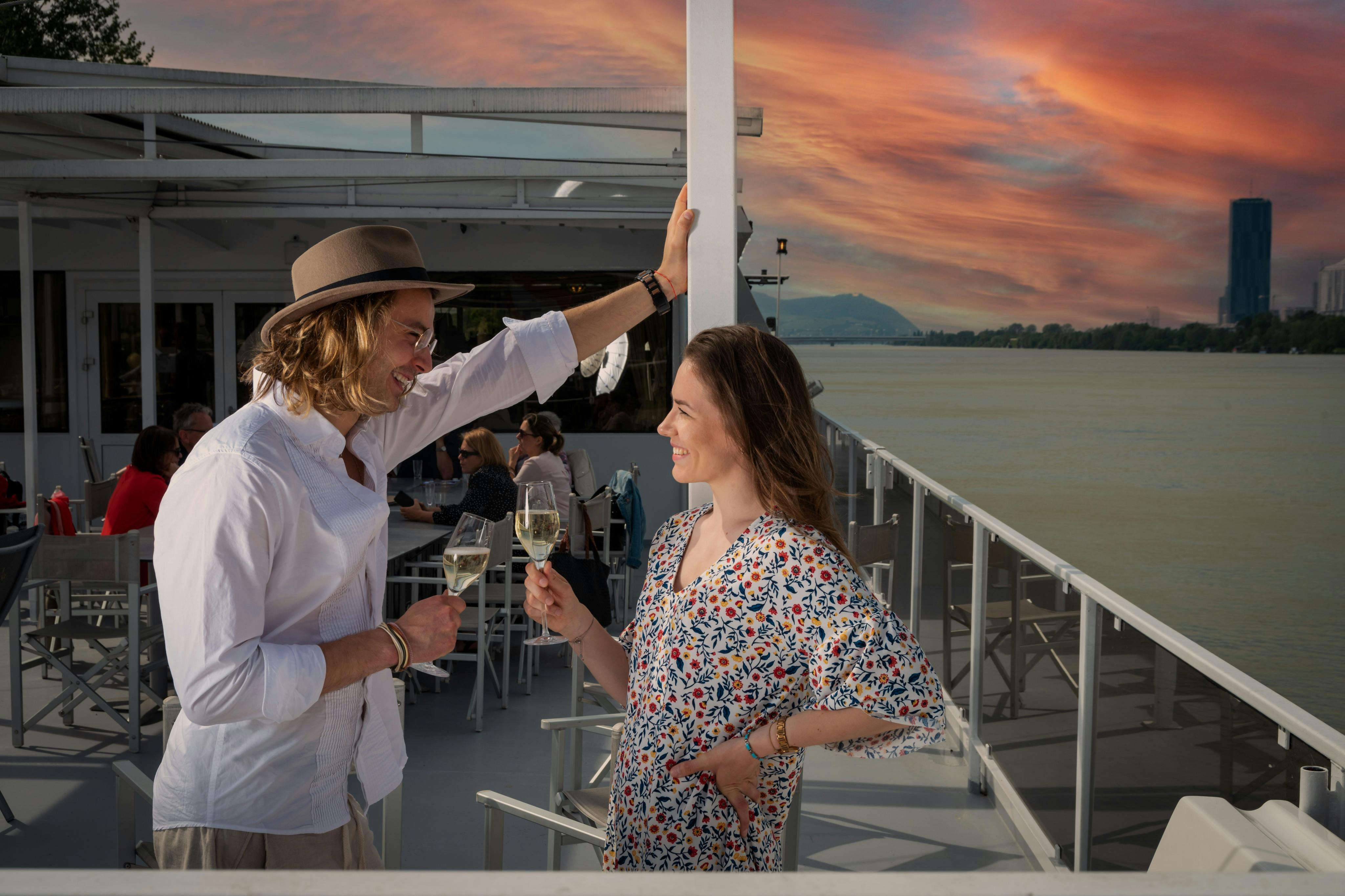 A man and woman clink glasses on a boat deck during sunset, with other people seated in the background.