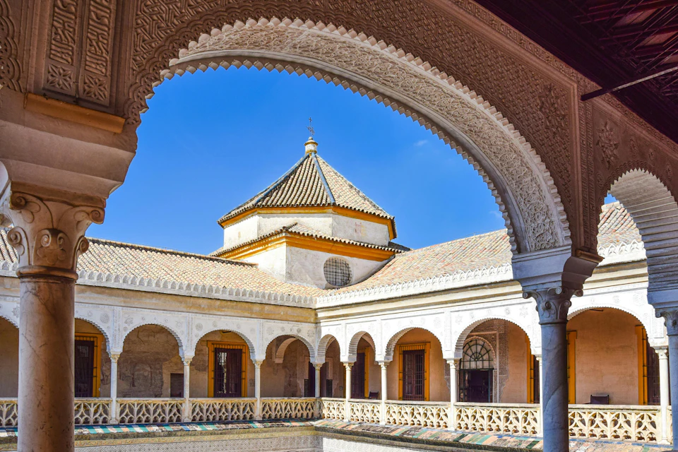 A courtyard with a central building featuring a tiled roof, surrounded by arched colonnades and ornate architectural details.