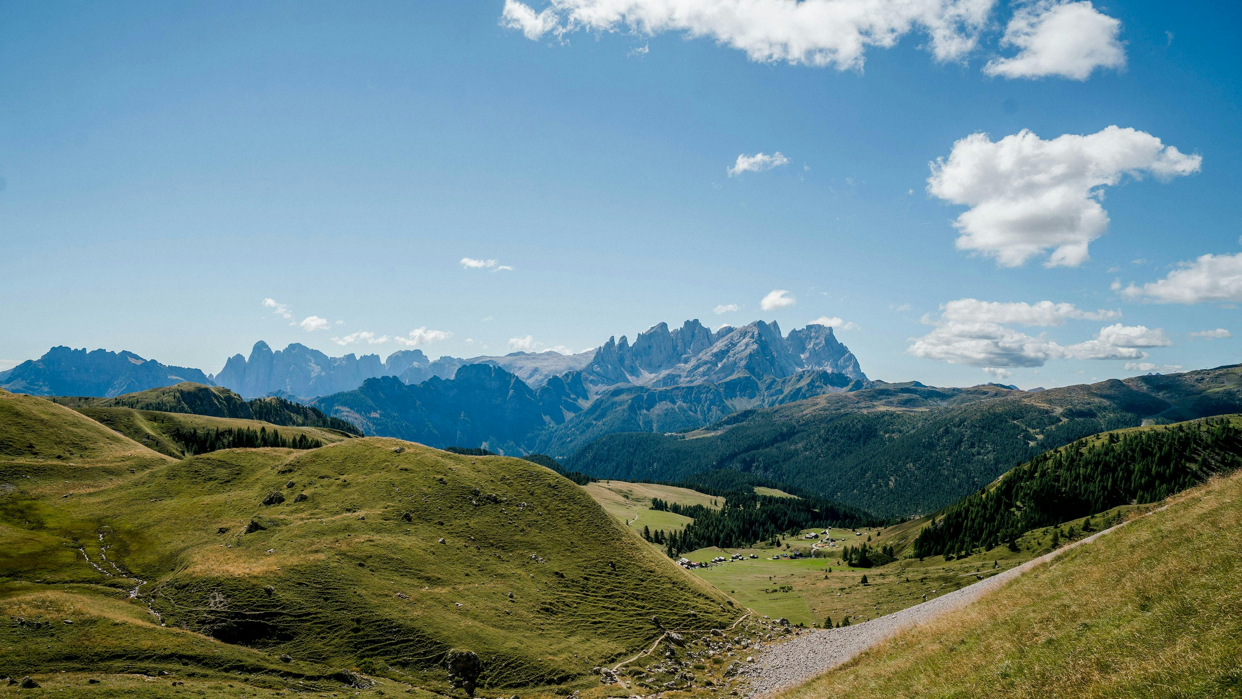Horská krajina se zelenými zvlněnými kopci, vzdálenými zubatými vrcholky, rozptýlenými mraky a malou vesnicí v údolí.