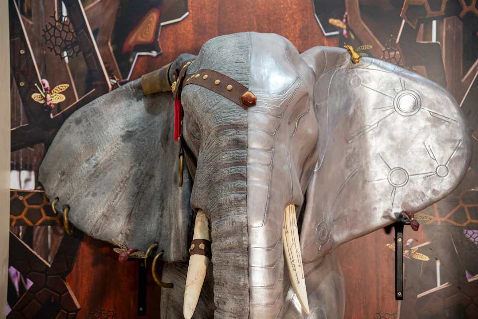 A sculptural display of an elephant's head with one half metallic and the other textured, featuring tusks and straps.