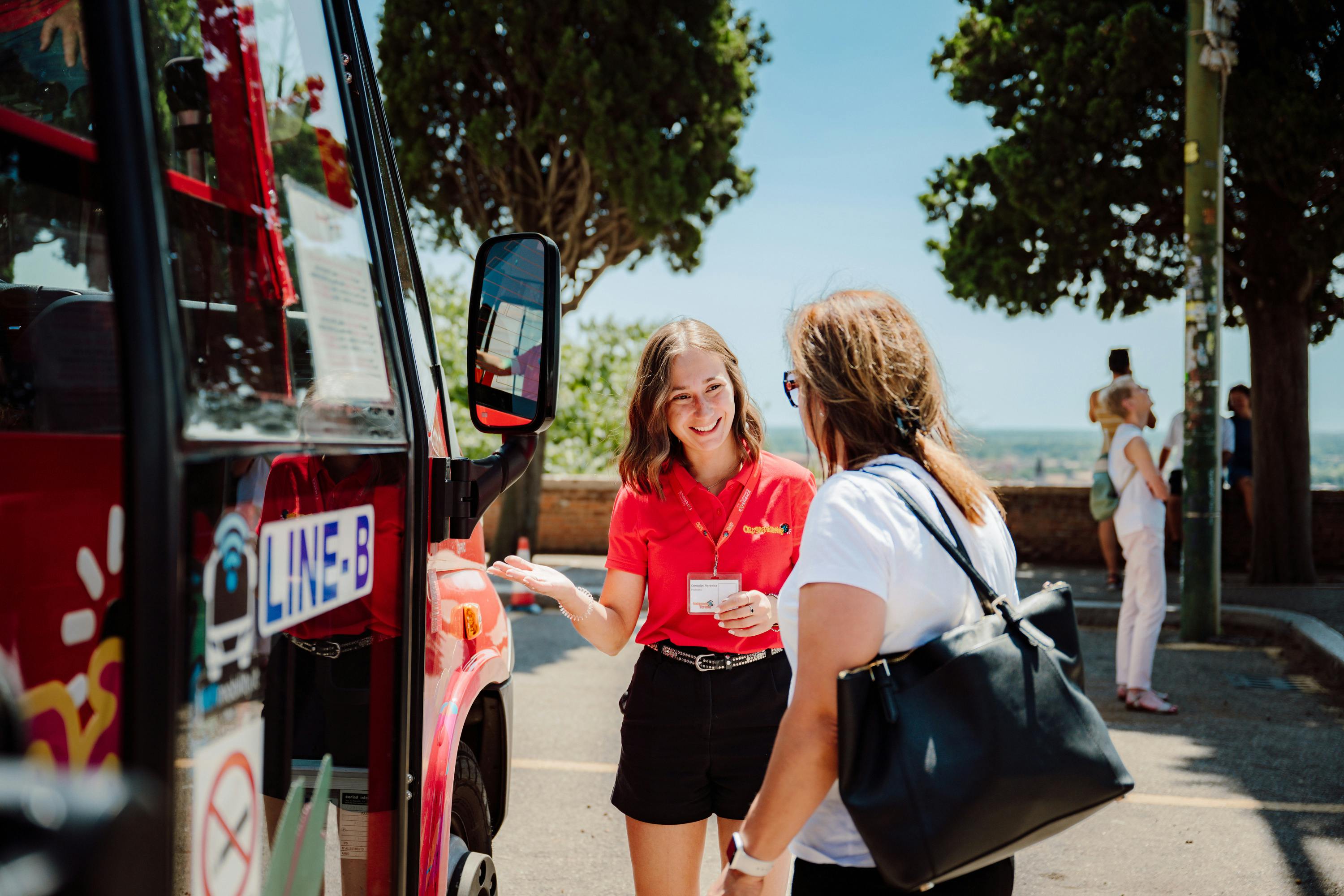 Una donna con una maglietta rossa interagisce con un'altra donna vicino a un autobus con la scritta 