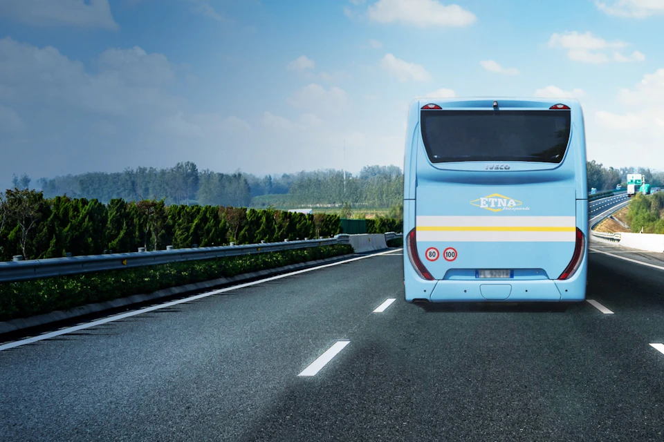 A blue bus travels on an empty highway surrounded by greenery under a partly cloudy sky.