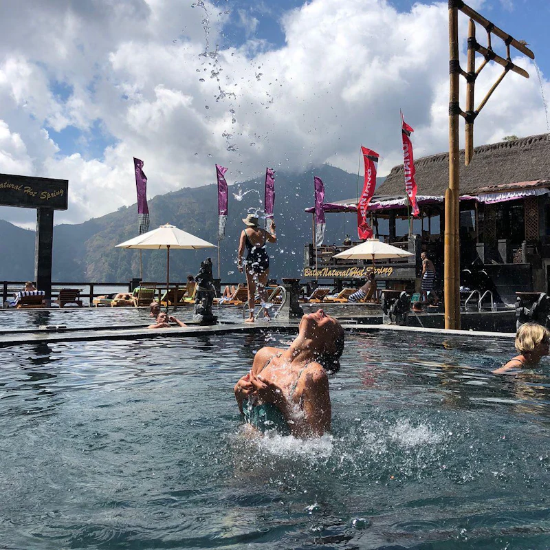 A person splashes water in a pool with mountains and a resort in the background under a partly cloudy sky.