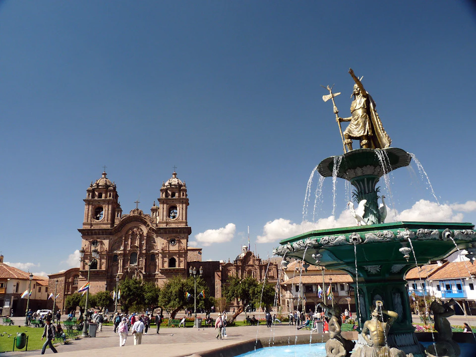 Historic cathedral with twin bell towers in the background, large ornate fountain with statue in the foreground, people walking.