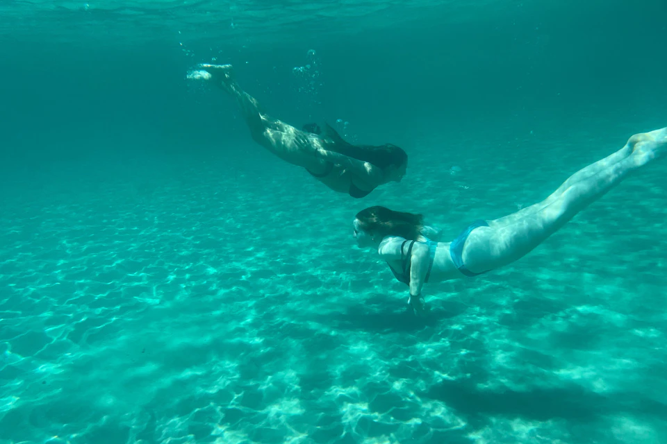 Two people swimming underwater in clear, turquoise water with sunlight illuminating the sandy bottom.