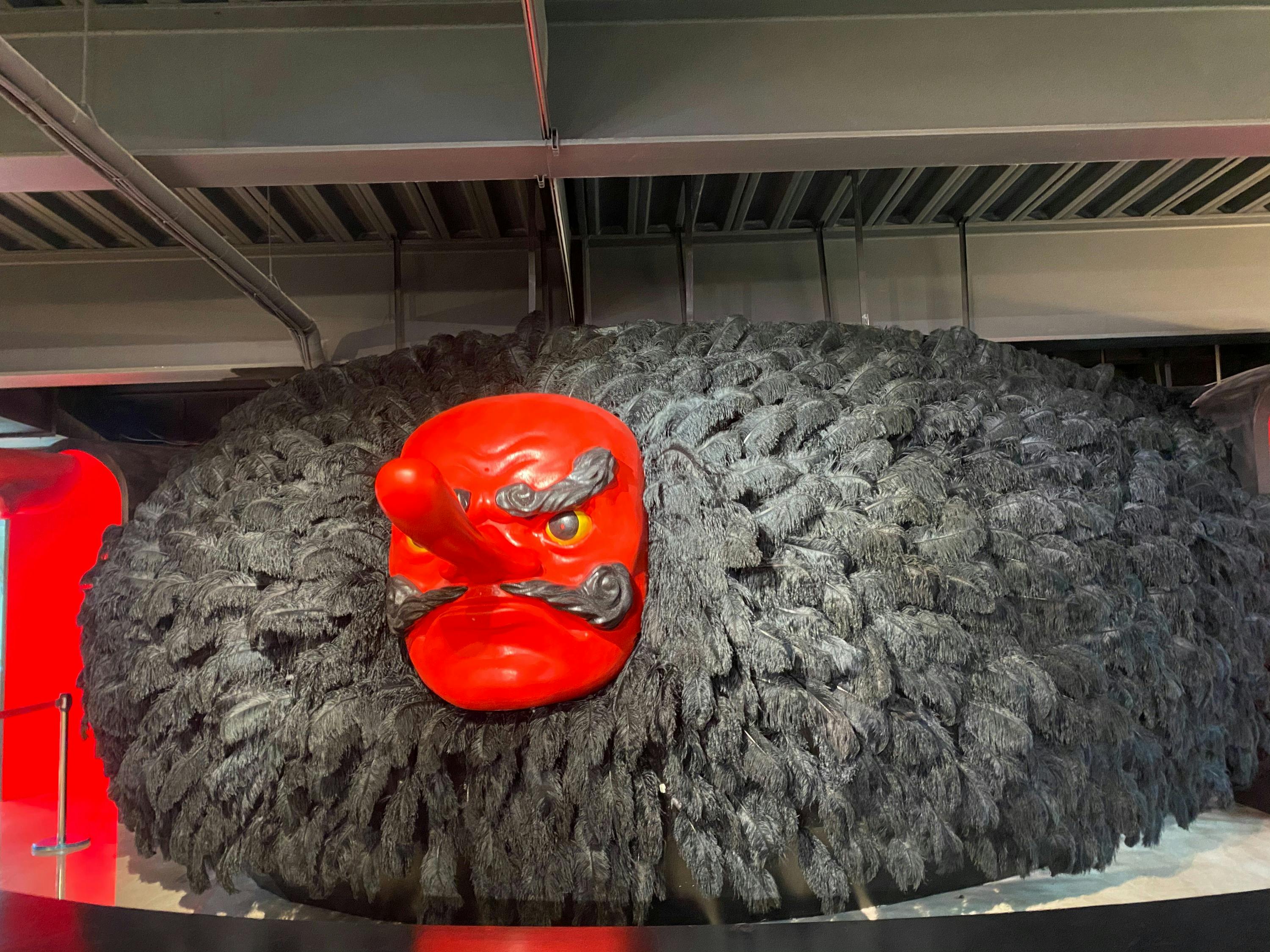 A large sculpture featuring a red mask with a long nose surrounded by black feathers, displayed indoors.