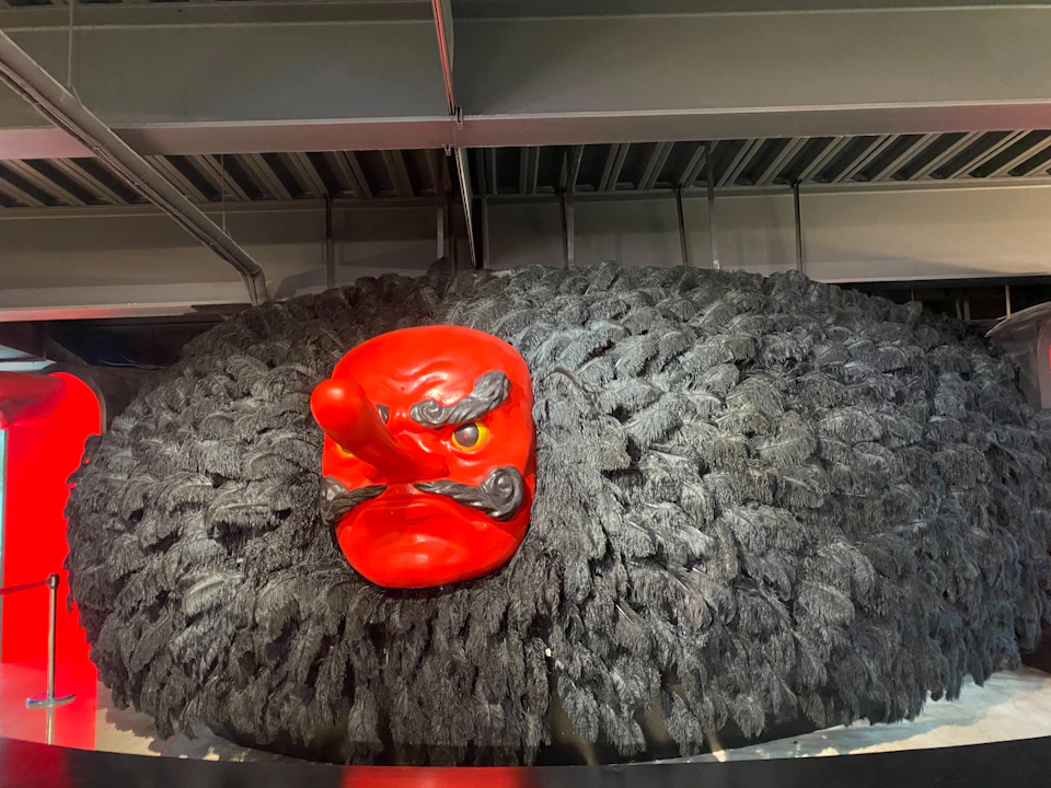 A red mask with a long nose and fierce expression is mounted on a textured black surface under industrial ceiling beams.