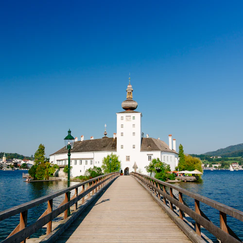 En lång träbro leder till en vit byggnad med en kupol, belägen på en sjö mot en kuliss av kullar och en klarblå himmel.