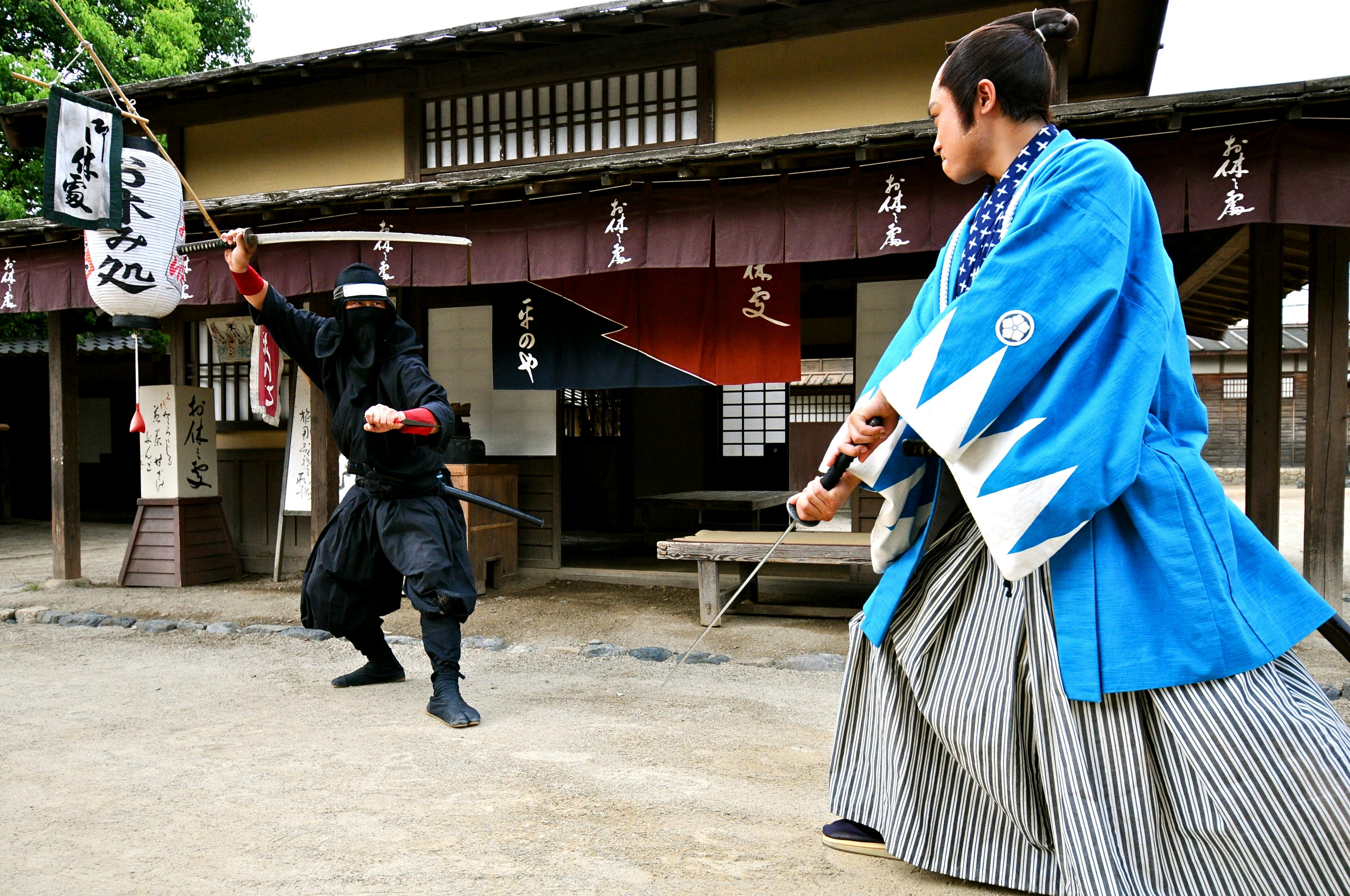 Eine Person in traditioneller japanischer Kleidung schwingt ein Schwert auf einem Feldweg vor einem traditionellen Gebäude mit hängenden Bannern.