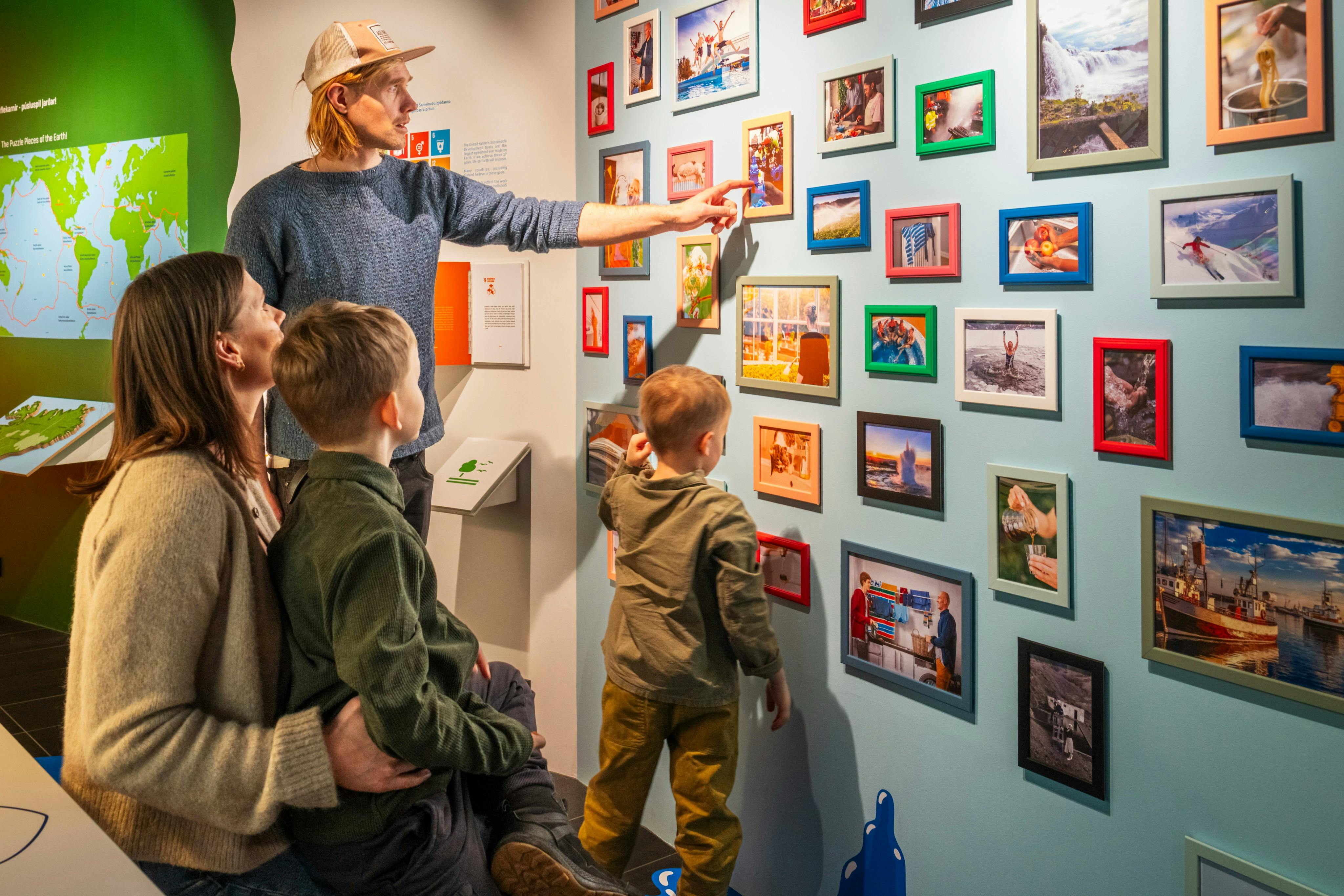 Eine vierköpfige Familie begutachtet eine Galeriewand mit verschiedenen gerahmten Bildern. Im Hintergrund sind eine Landkarte und ein Display zu sehen.