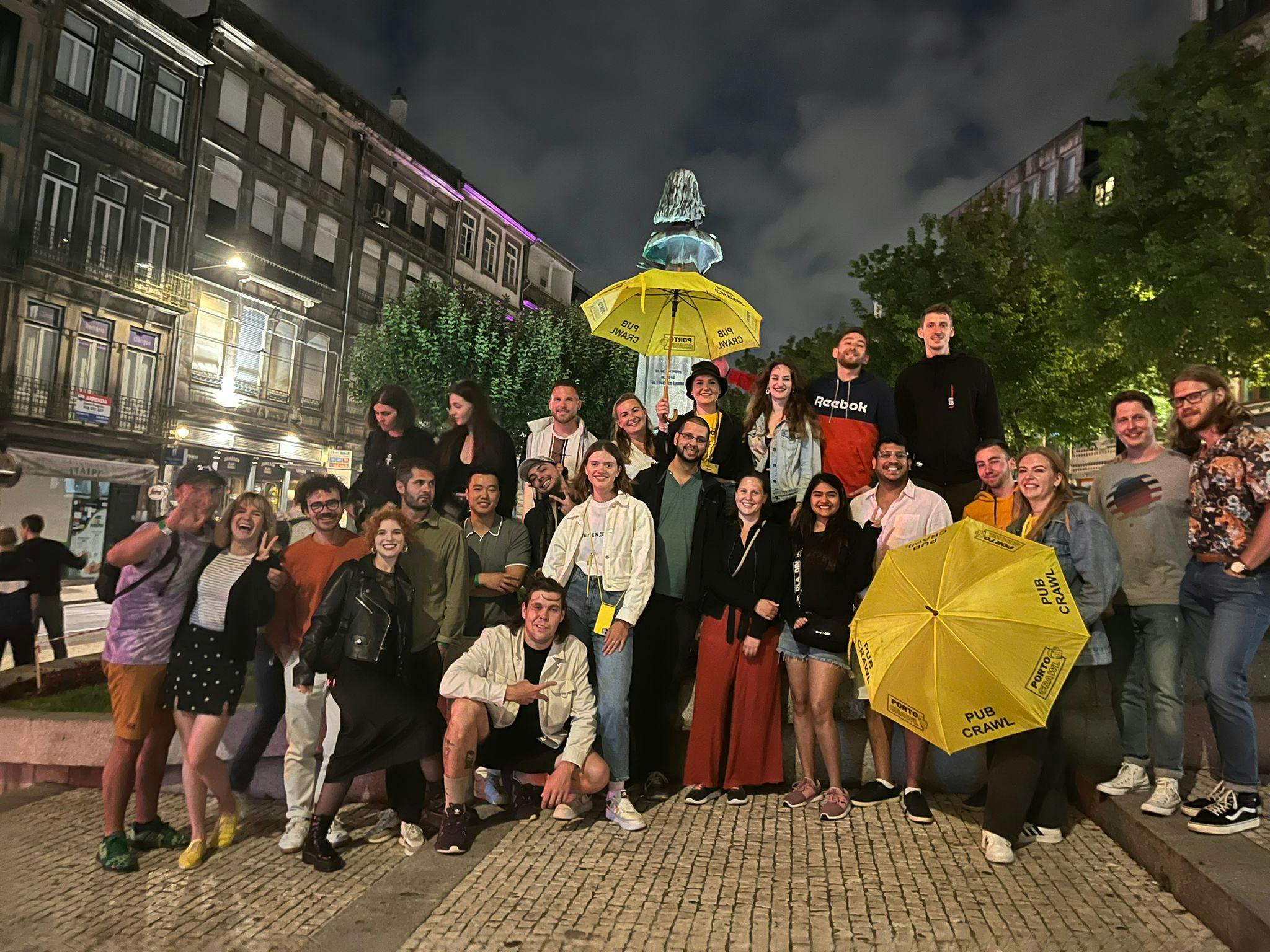A group of people posing at night in a city square, holding two yellow "Pub Crawl" umbrellas, with buildings and trees in the background.