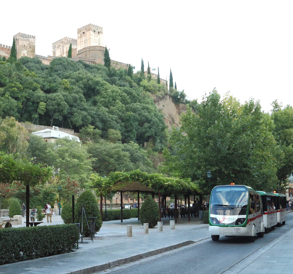 A white tour bus drives on a tree-lined road with a historic castle on a hill in the background.