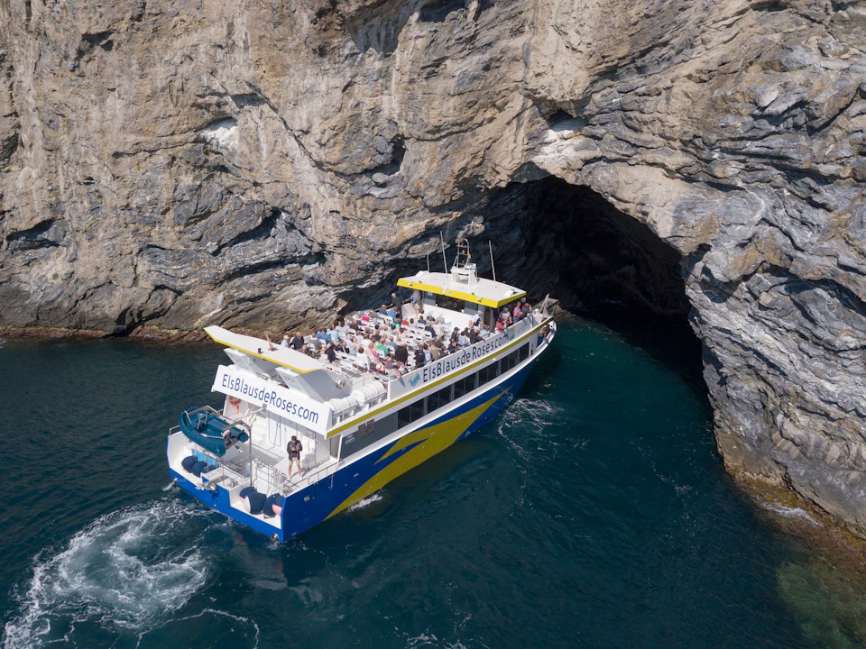 A boat full of passengers sails near a rocky cave entrance along a rugged coastline.