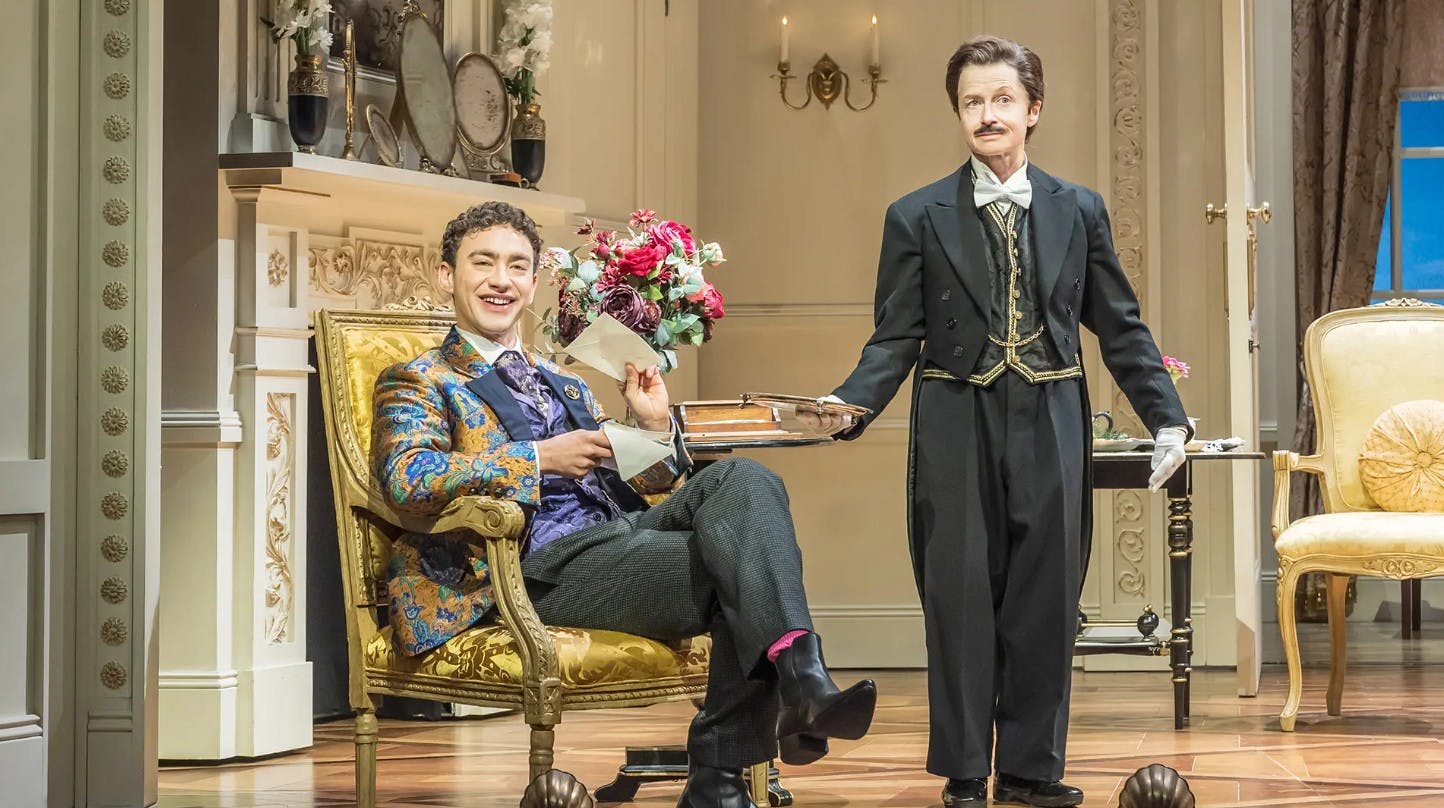 Δύο άνδρες με ενδυμασία του 19ου αιώνα αλληλεπιδρούν σε ένα περίτεχνο δωμάτιο με φλοράλ διαρρύθμιση και διακόσμηση αντίκα. Ο ένας είναι καθισμένος και χαμογελαστός, ο άλλος στέκεται.