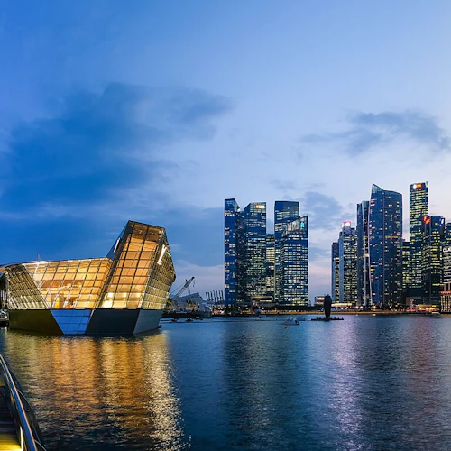 Moderne Skyline am Wasser in der Abenddämmerung mit beleuchteten Wolkenkratzern, einem markanten Gebäude in Form eines Bootes und einer geschwungenen Uferpromenade.