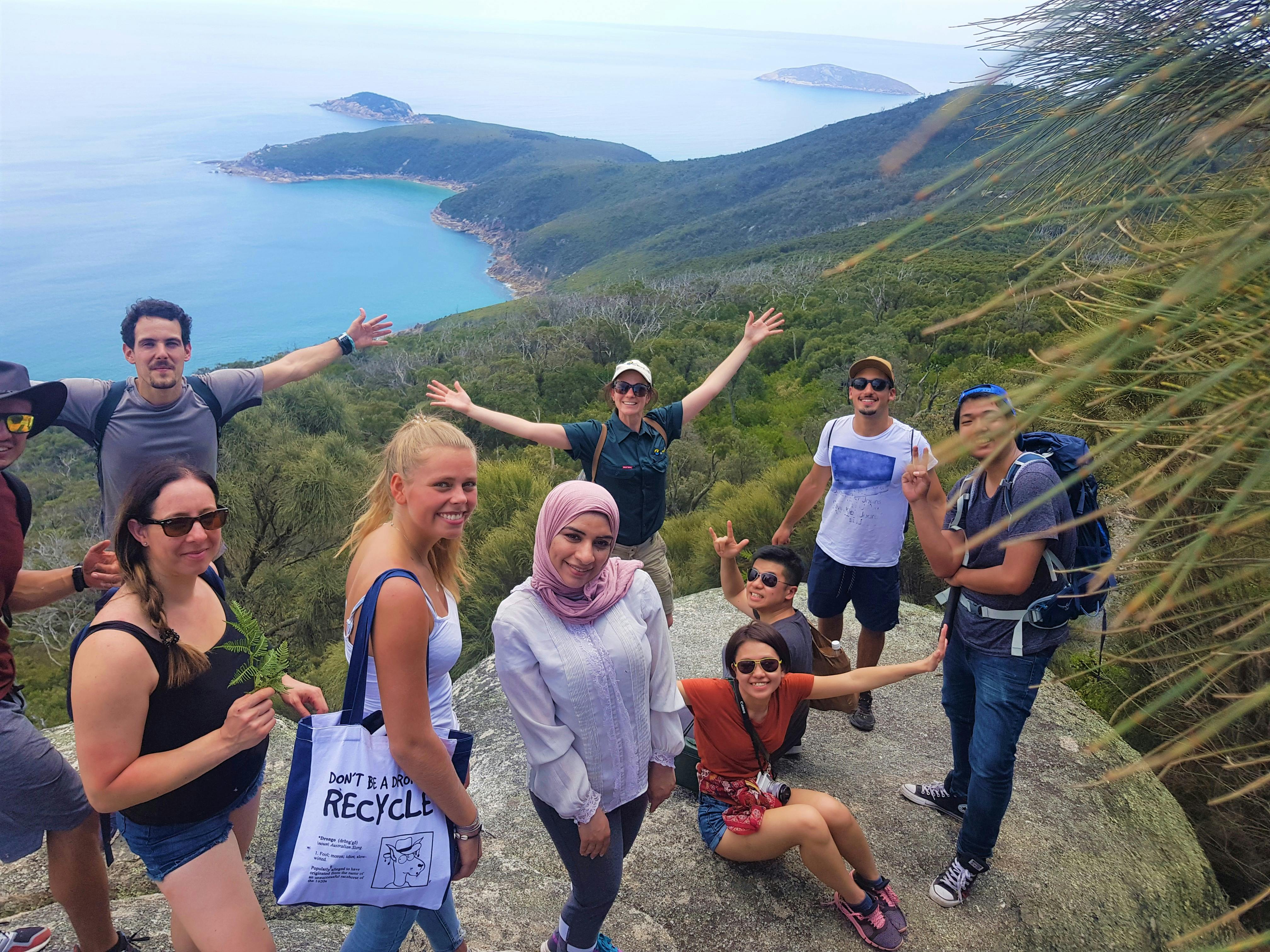 Μια ομάδα ανθρώπων που ποζάρουν σε ένα βράχο με γραφική θέα στην ακτογραμμή και τα νησιά, περιτριγυρισμένοι από πράσινο και καταγάλανα νερά.