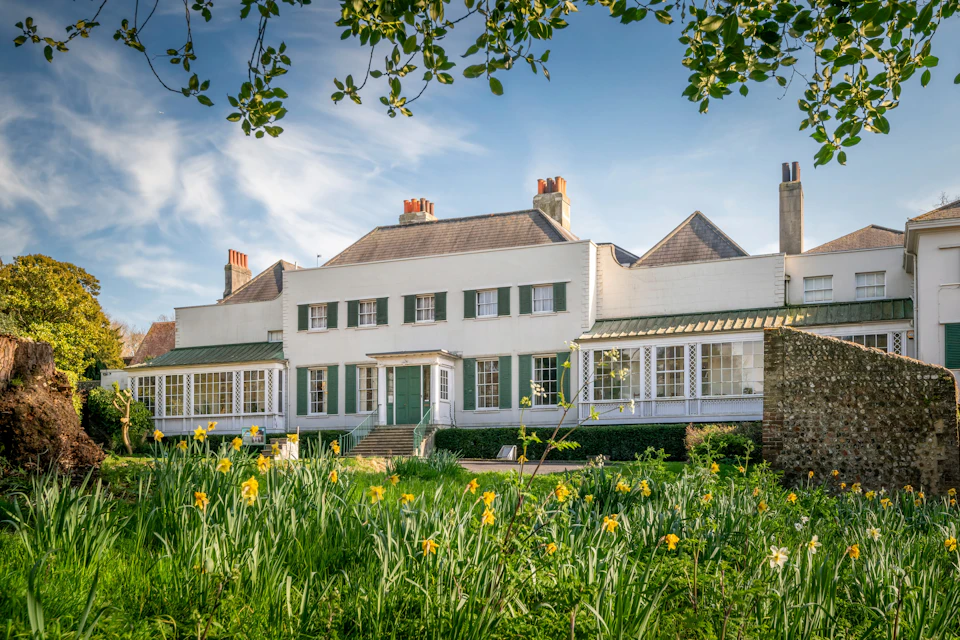 A white, two-story house with green shutters, surrounded by blooming flowers and greenery, viewed from the garden.