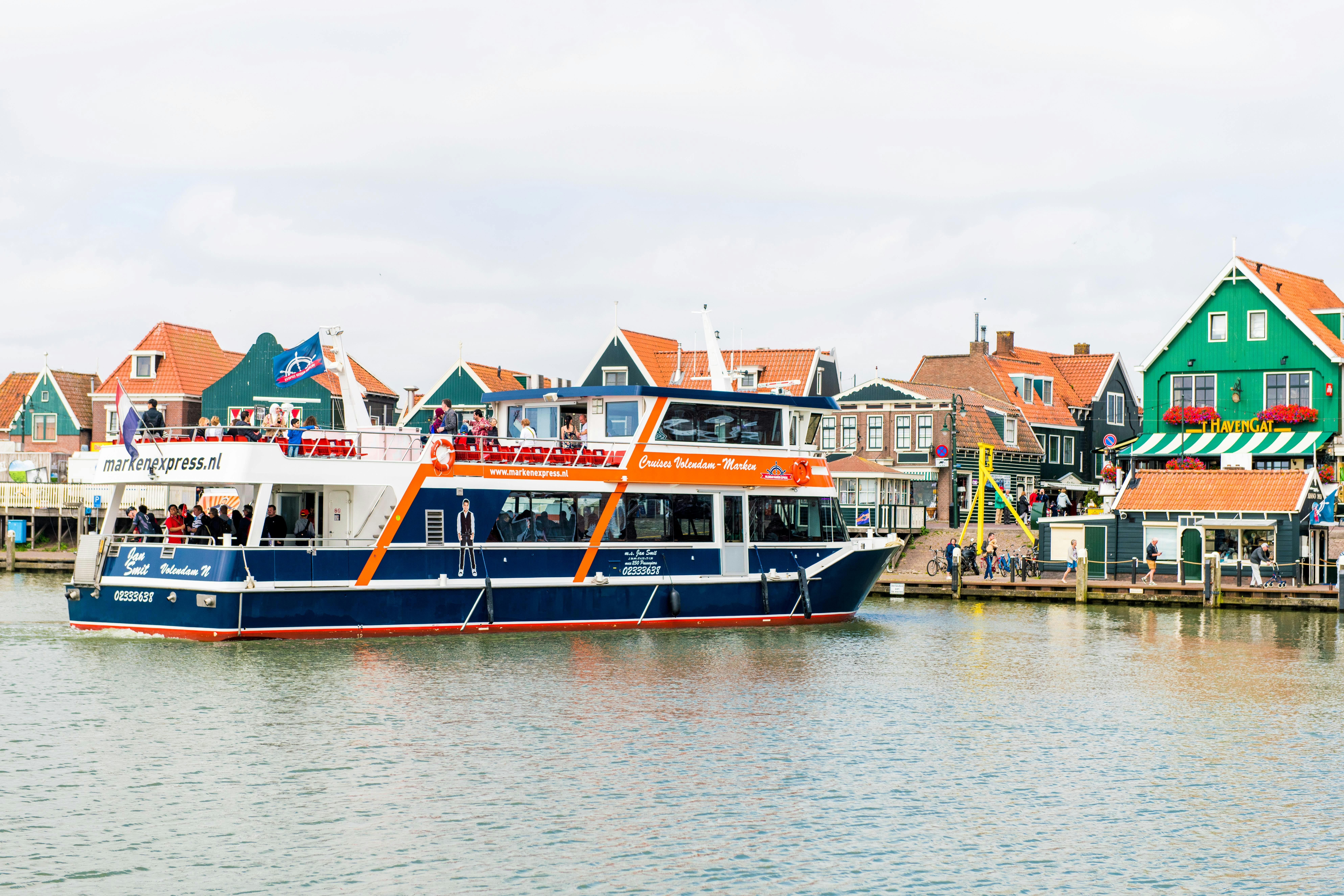 Ένα κρουαζιερόπλοιο δύο επιπέδων με επιβαίνοντες είναι αγκυροβολημένο κοντά σε μια ακτή με παραδοσιακά ευρωπαϊκά κτίρια.