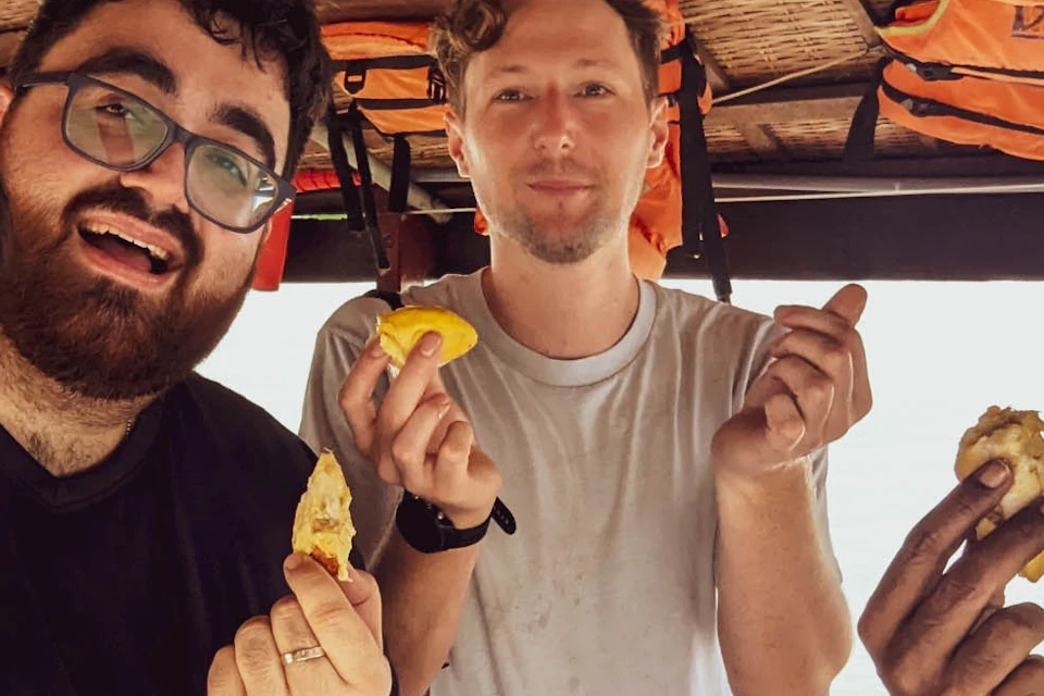 Four men joyfully holding pieces of food on a wooden boat, with life jackets hanging above them.