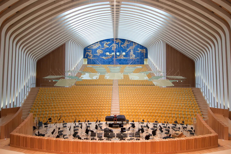 Empty orchestral concert hall with rows of yellow seats, a grand piano on stage, and artistic murals above the seating area.