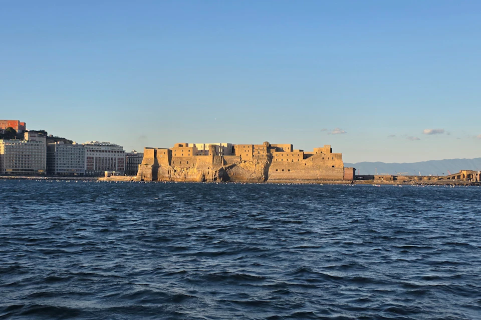 A historic stone castle on a rocky base surrounded by water, under a clear blue sky, with a cityscape in the background.