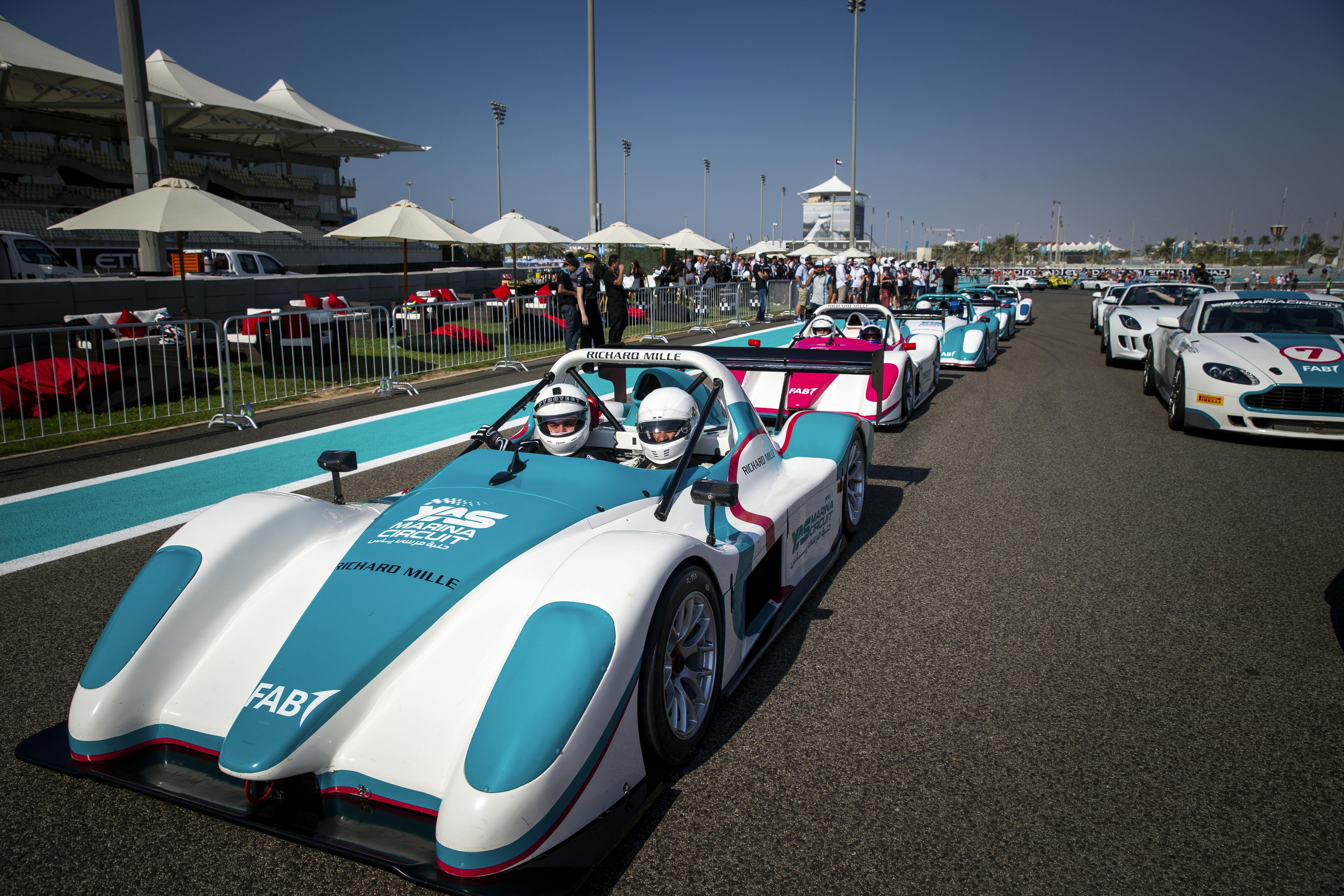 Race cars lined up on a track with drivers wearing helmets. Spectators and umbrellas are visible in the background.
