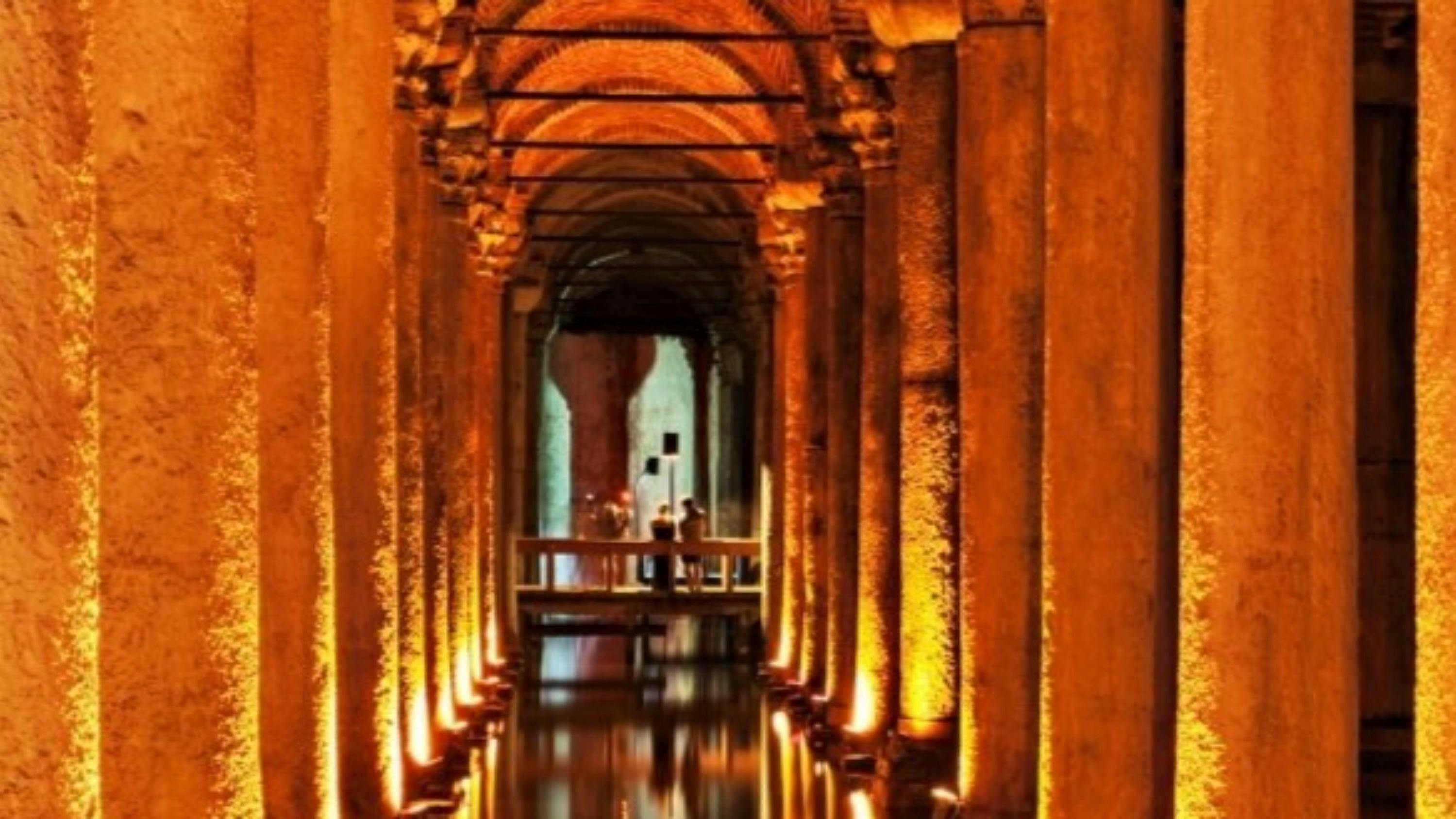 An ancient underground columned hall with warm lighting, a reflective water surface, and a bridge with people in the distance.