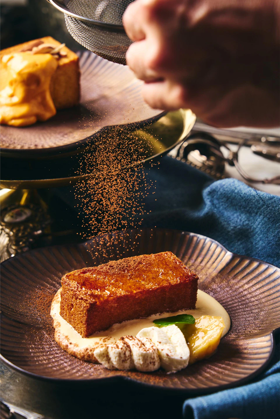 A plated dessert with a piece of cake, cream, and garnish, being dusted with cocoa powder by a hand.