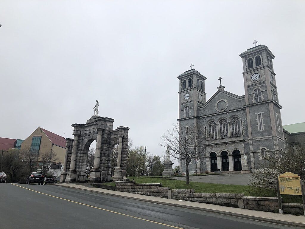 흐린 날에는 쌍둥이 시계탑이 있는 회색 석조 교회가 석조 아치 기념비 옆에 서 있으며, 근처에 주차된 차량이 몇 대 있습니다.