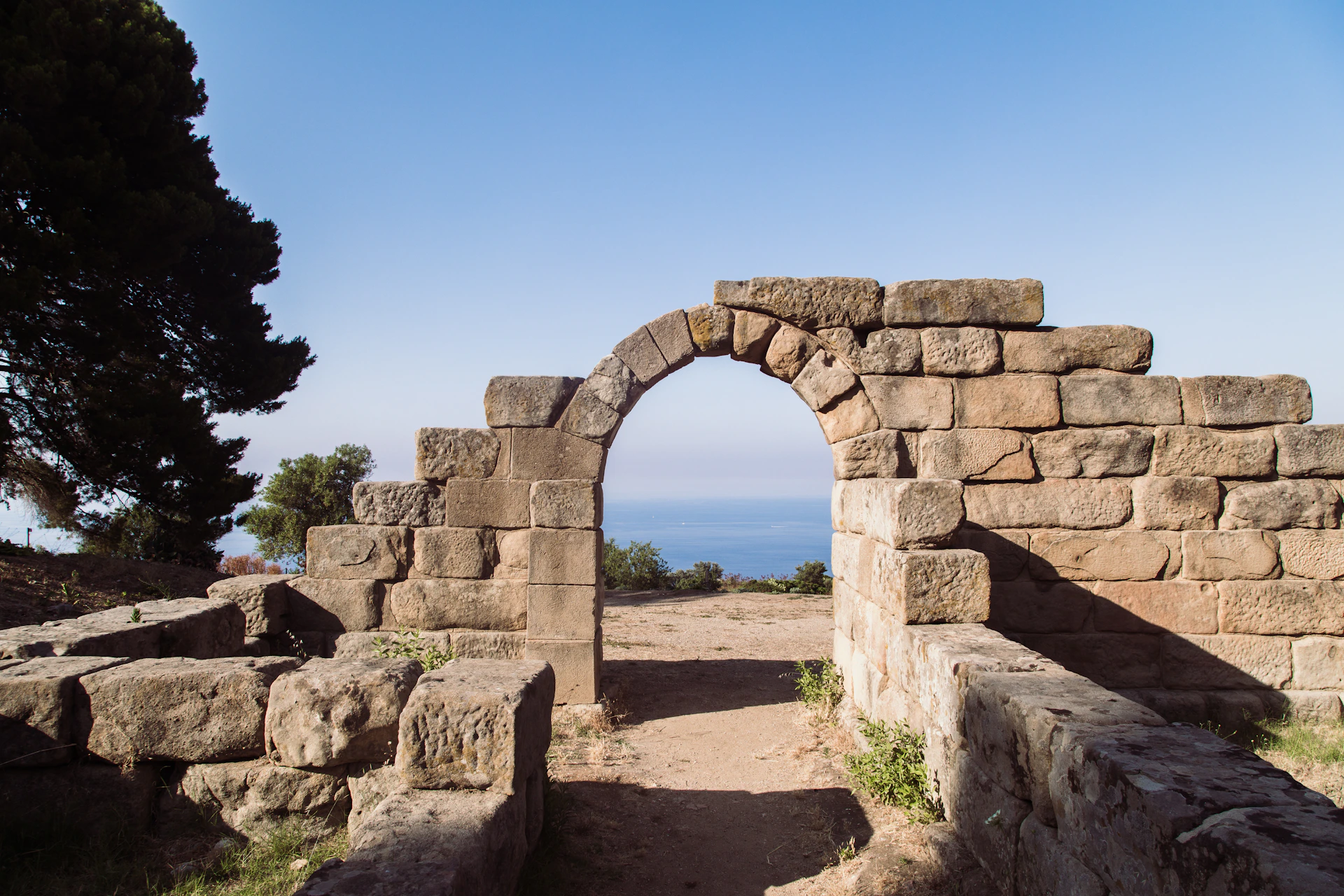 Stone archway ruins with a dirt path leading to a view of the sea under a clear blue sky. Trees and shrubbery are visible.
