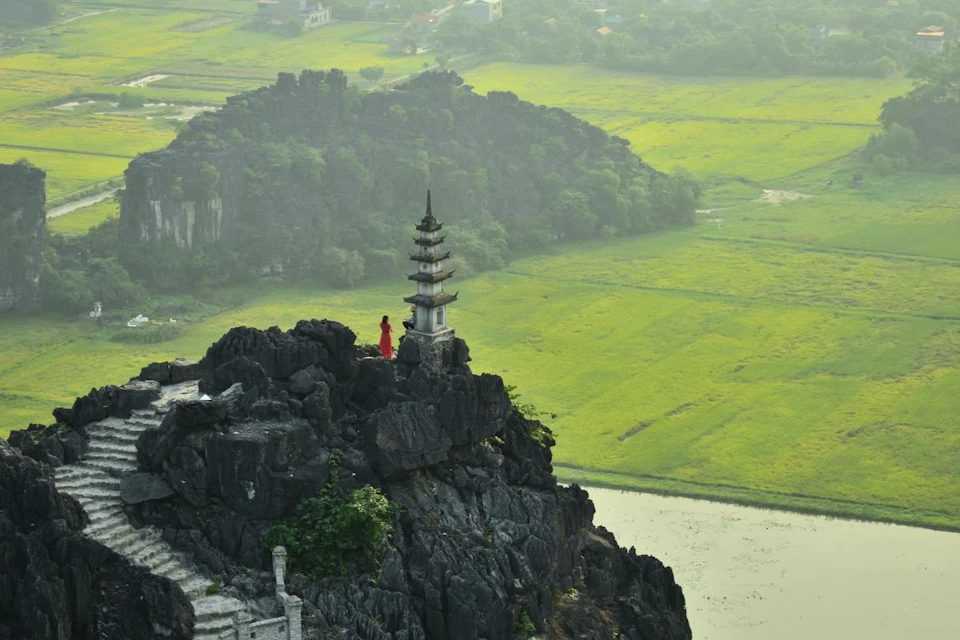 A person in a red dress stands on a rocky mountain ledge beside a pagoda, overlooking expansive green fields.