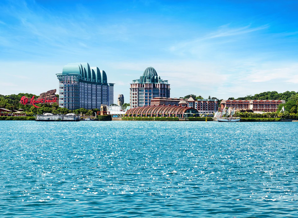 Buildings with distinctive domed roofs and a waterfront are set against a blue sky, with sparkling water in the foreground.