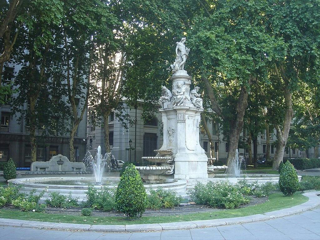 Kamienna fontanna z ozdobnymi rzeźbami, otoczona strumieniami wody, zielenią i dużymi drzewami, osadzona na terenie parku miejskiego.