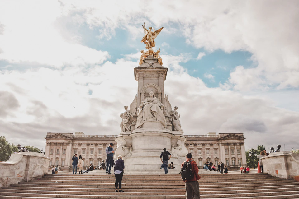 People walking around and taking photos near the Victoria Memorial in front of Buckingham Palace under a partly cloudy sky.