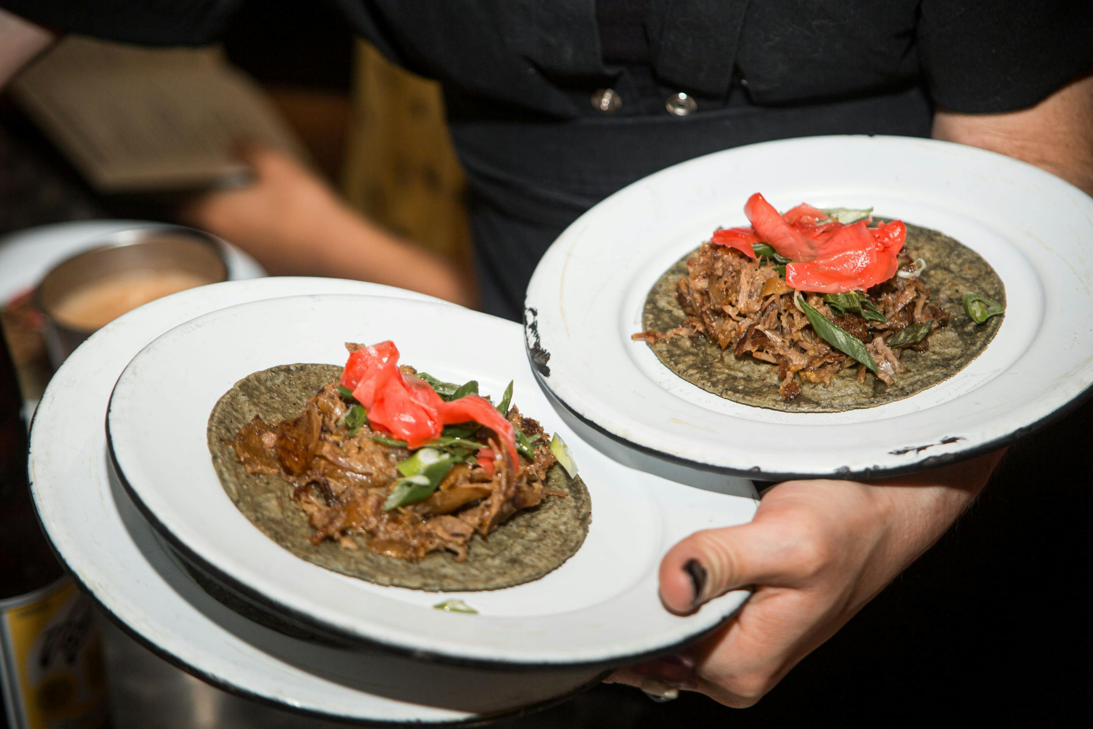 Une personne tenant deux assiettes de tacos avec de la viande, des herbes et des garnitures rouges, présentés sur des tortillas foncées.
