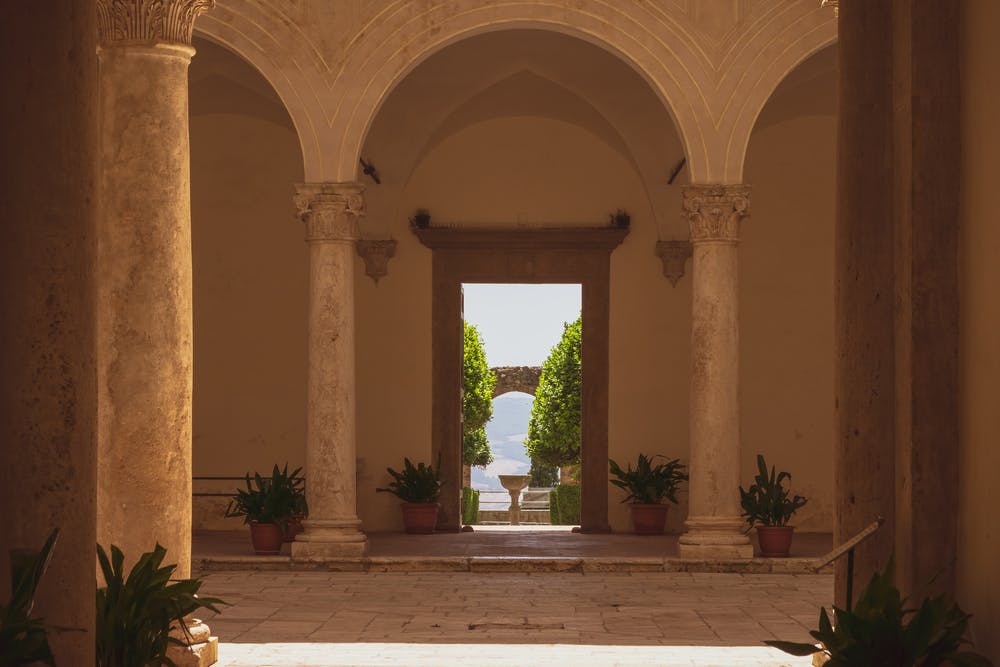 Τοξωτός διάδρομος με κίονες οδηγεί σε μια εξωτερική αυλή με γλάστρες, πράσινα δέντρα και μια μακρινή πέτρινη αψίδα.