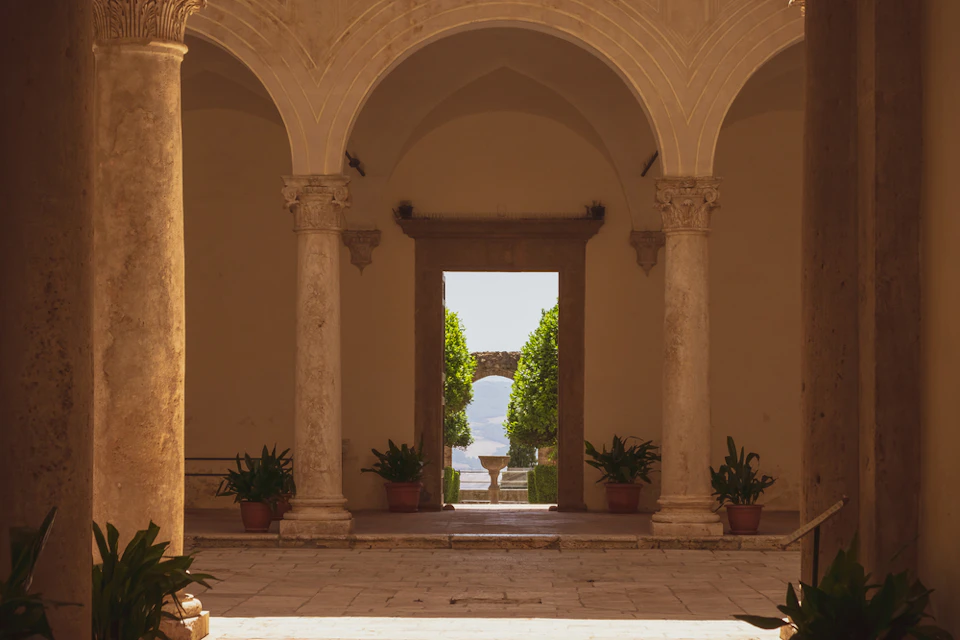 Arched hallway with columns leads to an outdoor courtyard featuring potted plants, green trees, and a distant stone arch.