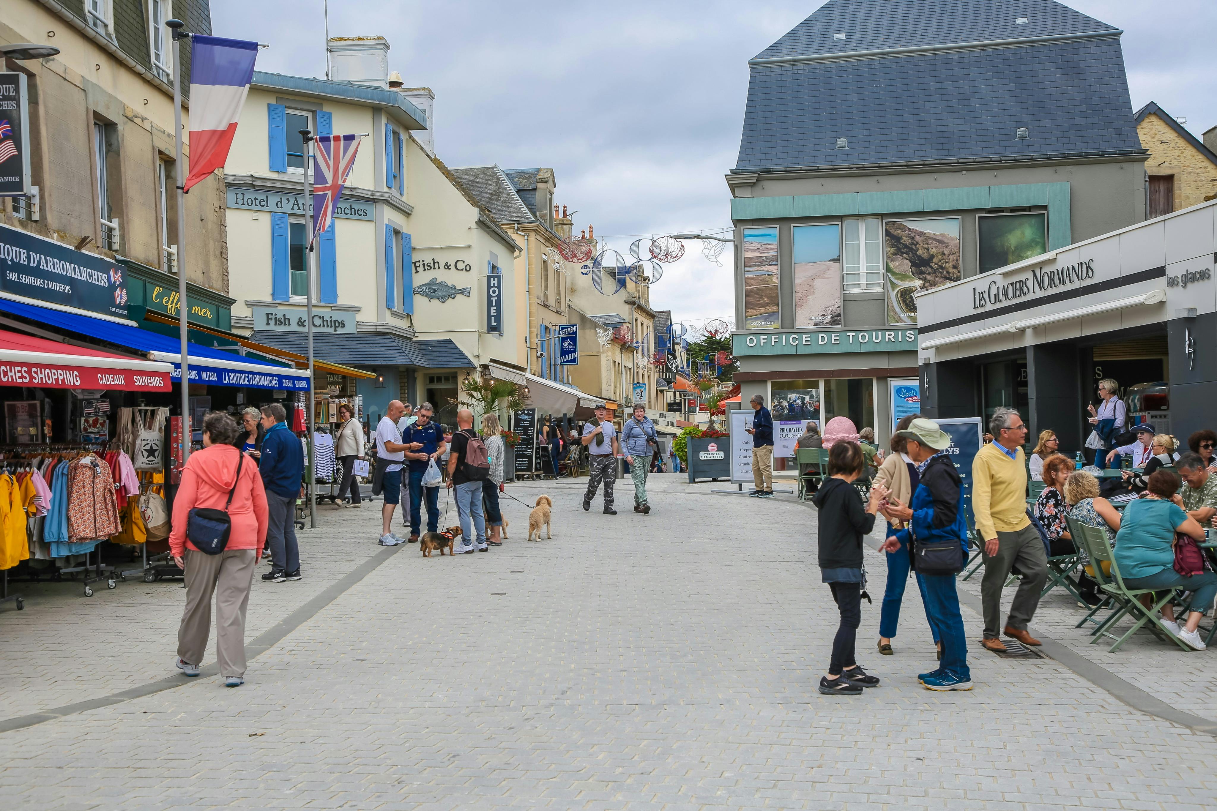 Οι άνθρωποι περπατούν και κάθονται σε ένα υπαίθριο καφέ σε έναν πεζόδρομο με καταστήματα και ένα τουριστικό γραφείο στο παρασκήνιο.