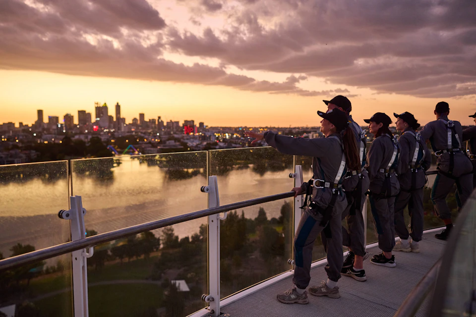 Group of people in harnesses stand on a platform overlooking a cityscape at sunset, with water and trees below.