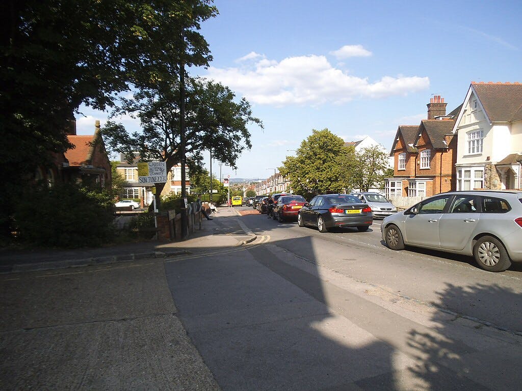 Une rue de banlieue bordée de voitures garées, de maisons en briques et d'arbres, sous un ciel bleu clair avec des nuages épars.