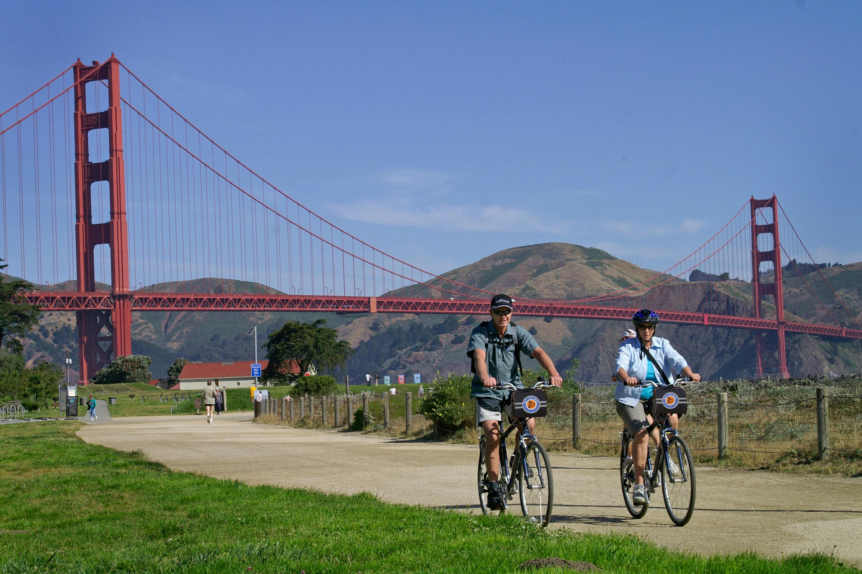 Two people biking on a path near greenery, with the Golden Gate Bridge and hills in the background under a clear blue sky.