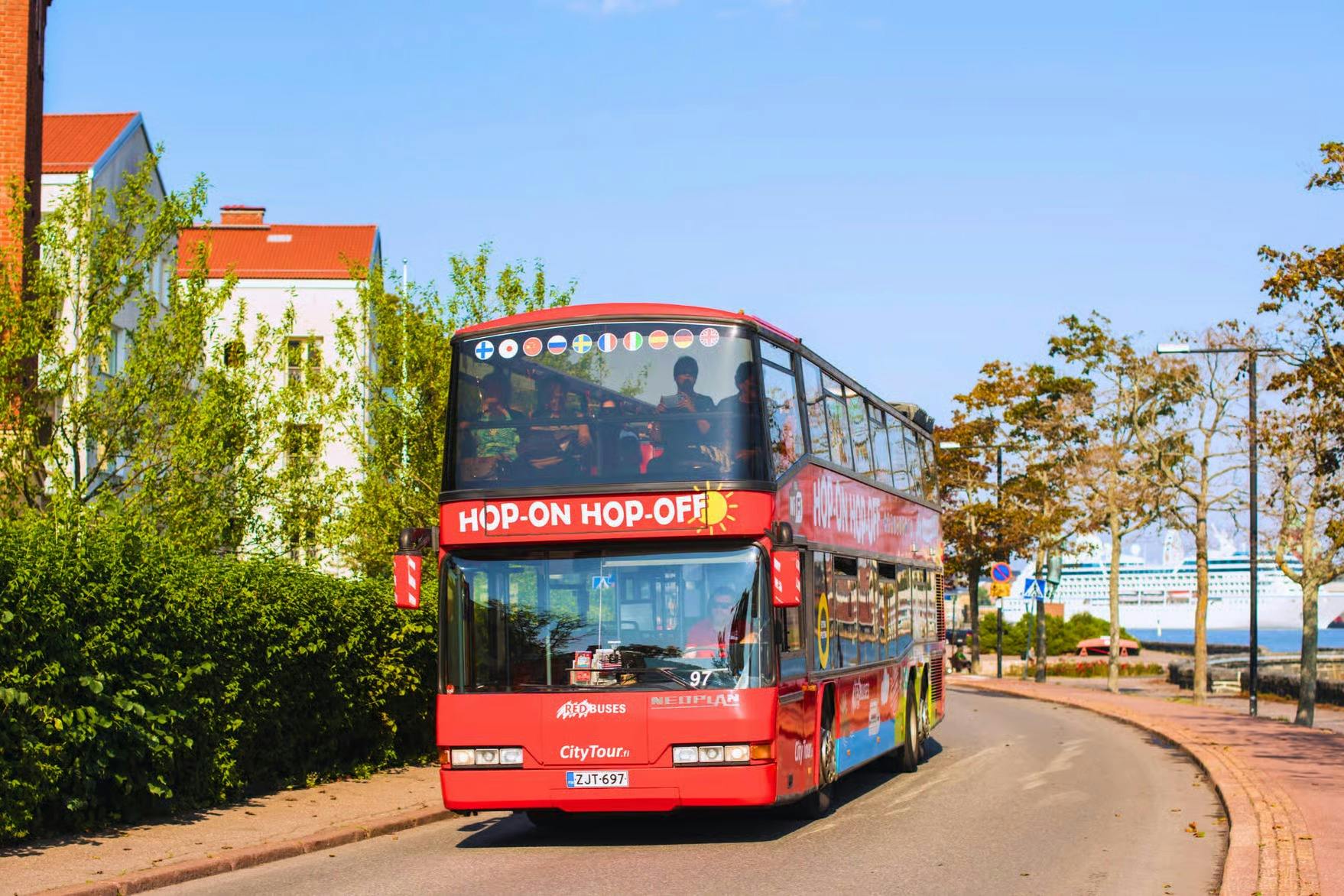 Červený dvoupatrový zájezdový autobus s označením „Hop-On Hop-Off“ jedoucí po slunné ulici obklopené zelení a budovami.