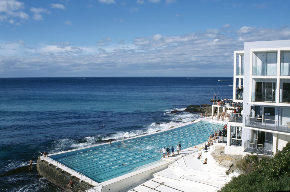 A beachfront swimming pool with people swimming and others standing nearby, next to a building and overlooking the ocean.