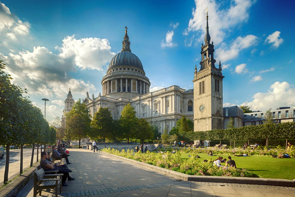 A large cathedral with a dome and towers surrounded by a park with benches, trees, and people enjoying the scenery.