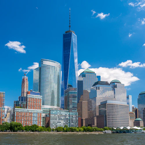 Mehrere moderne Wolkenkratzer, darunter ein hoher, spitzer Turm, stehen entlang der Uferpromenade unter einem klaren blauen Himmel.