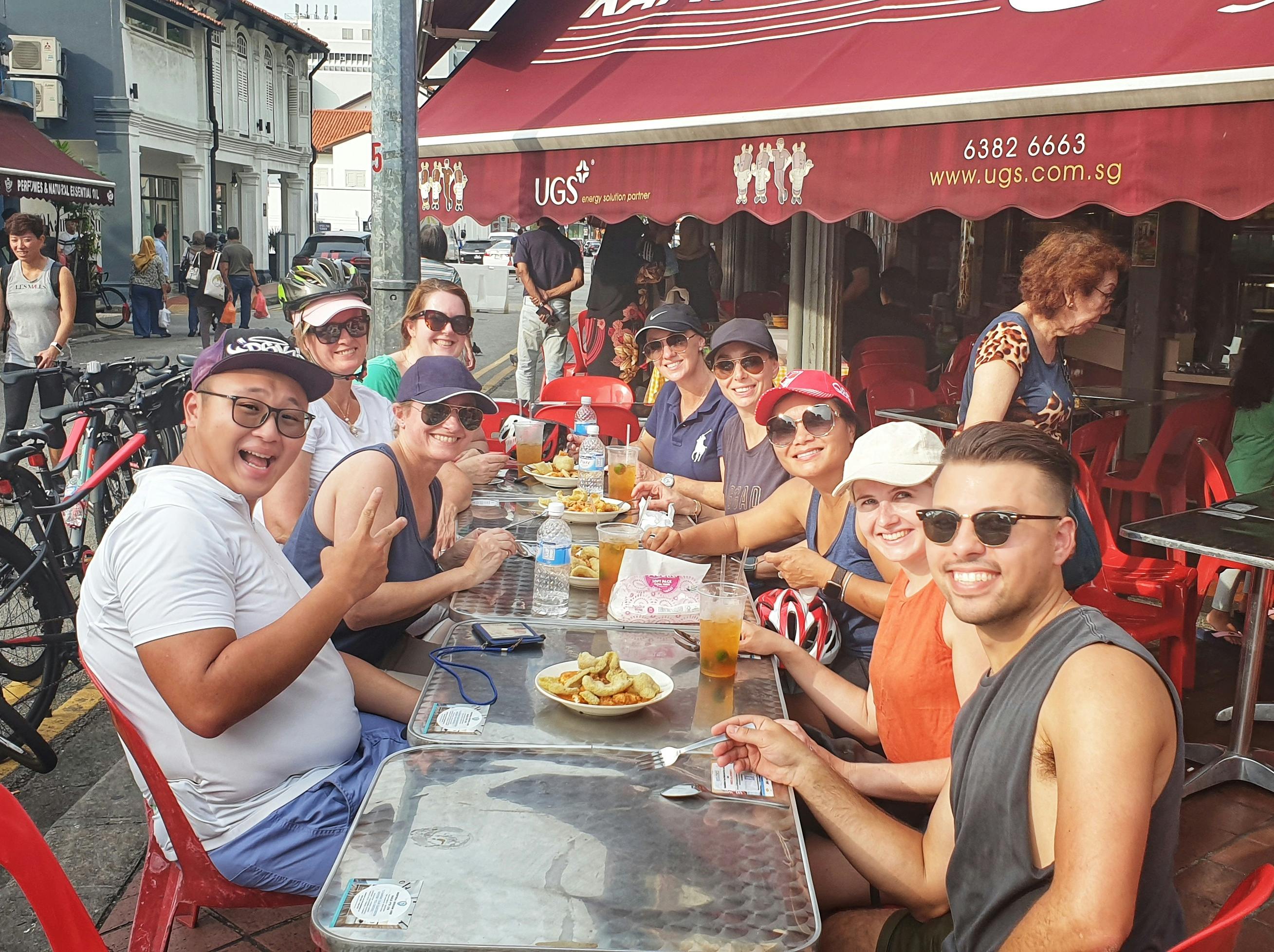 En gruppe smilende mennesker, der sidder ved et udendørs cafébord og nyder mad og drikkevarer under en rød baldakin.