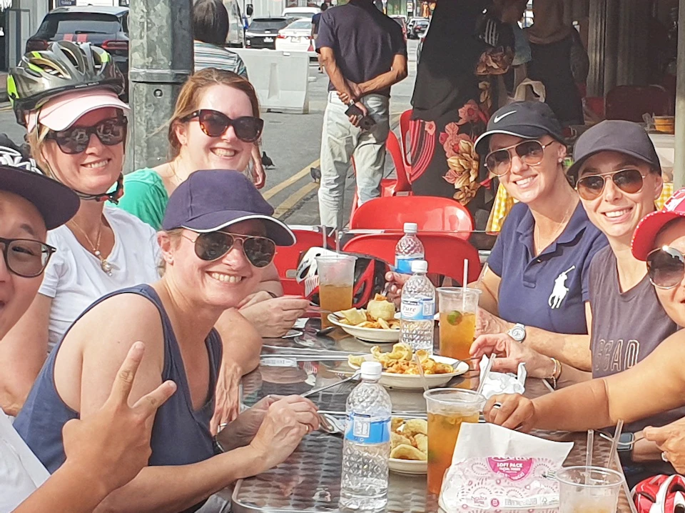 A group of smiling people sitting at an outdoor café table, enjoying food and drinks under a red canopy.