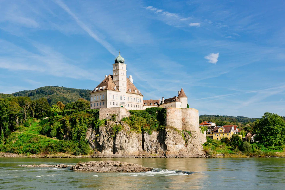 A large castle with a green dome sits on a rocky cliff overlooking a river, with a village and forested hills in the background.