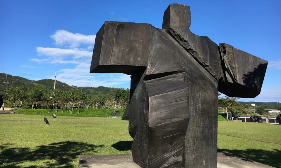 A large abstract black sculpture resembling a human figure in a sunny park with grass, trees, and a blue sky.