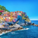 Colorful cliffside houses overlook the blue sea, with boats on rocky shore and greenery on the hillside under a clear sky.